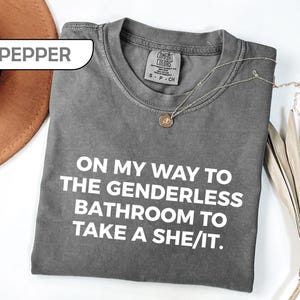 May include: A pepper-colored crew neck t-shirt with white text that reads ON MY WAY TO THE GENDERLESS BATHROOM TO TAKE A SHE/IT. The shirt features a Comfort Colors brand label at the inner neckline and is displayed with a gold-toned necklace.