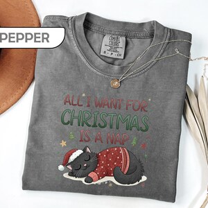 May include: A pepper-colored t-shirt featuring a graphic of a black cat wearing a red sweater and a Santa hat. The text reads "ALL I WANT FOR CHRISTMAS IS A NAP" in red and green letters. The shirt has a crew neckline and a tag labeled "PEPPER".