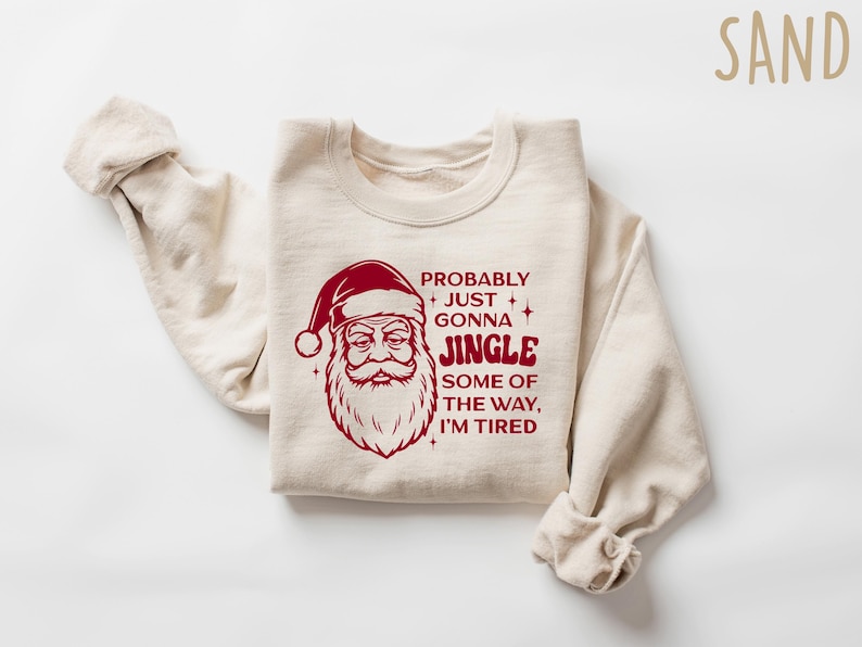 May include: Sand-colored sweatshirt with a red Santa Claus graphic and the text "Probably Just Gonna Jingle Some of the Way, I'm Tired." The long-sleeved top is folded on a white surface.