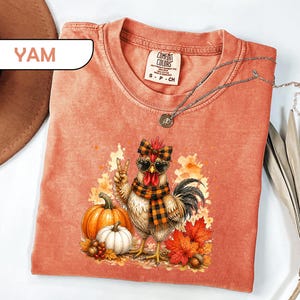 May include: A coral-colored t-shirt featuring a graphic of a chicken wearing a plaid scarf, a matching bow, and heart-shaped sunglasses. The chicken is making a peace sign next to pumpkins, autumn leaves, and acorns. The shirt has a crew neck.