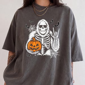 May include: A charcoal gray short-sleeve t-shirt featuring a white graphic of a skeleton yeti wearing sunglasses. The yeti holds an orange jack-o'-lantern pumpkin and makes a rock-and-roll hand gesture, surrounded by white spiderwebs and bats.