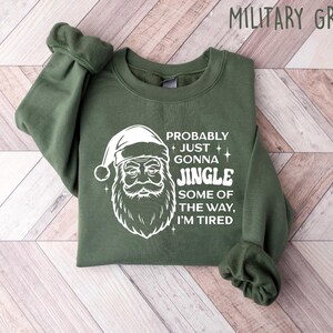 May include: Military green sweatshirt with a white Santa Claus graphic and the text "Probably Just Gonna Jingle Some of the Way, I'm Tired." The long-sleeved top is folded on a light wood-grain surface.