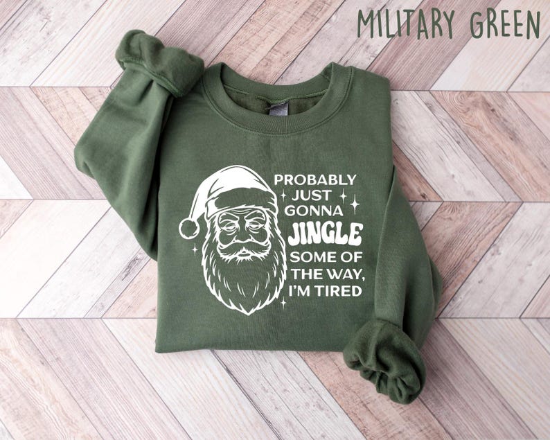 May include: Military green sweatshirt with a white Santa Claus graphic and the text "Probably Just Gonna Jingle Some of the Way, I'm Tired." The long-sleeved top is folded on a light wood-grain surface.