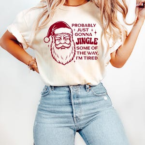 May include: Cream-colored t-shirt featuring a Santa Claus graphic in burgundy with the text "Probably Just Gonna Jingle Some of the Way, I'm Tired." The shirt is paired with light blue denim jeans.