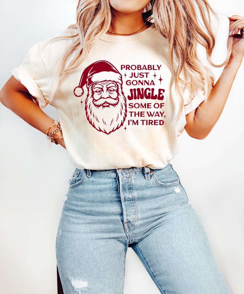 May include: Cream-colored t-shirt featuring a Santa Claus graphic in burgundy with the text "Probably Just Gonna Jingle Some of the Way, I'm Tired." The shirt is paired with light blue denim jeans.