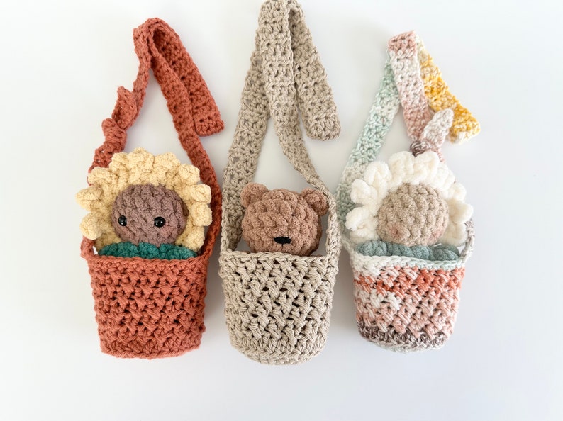 May include: Three crocheted bags with straps, each featuring a different crocheted animal inside. The bags are in shades of brown, beige, and pink. The animals are a sunflower, a bear, and a daisy.
