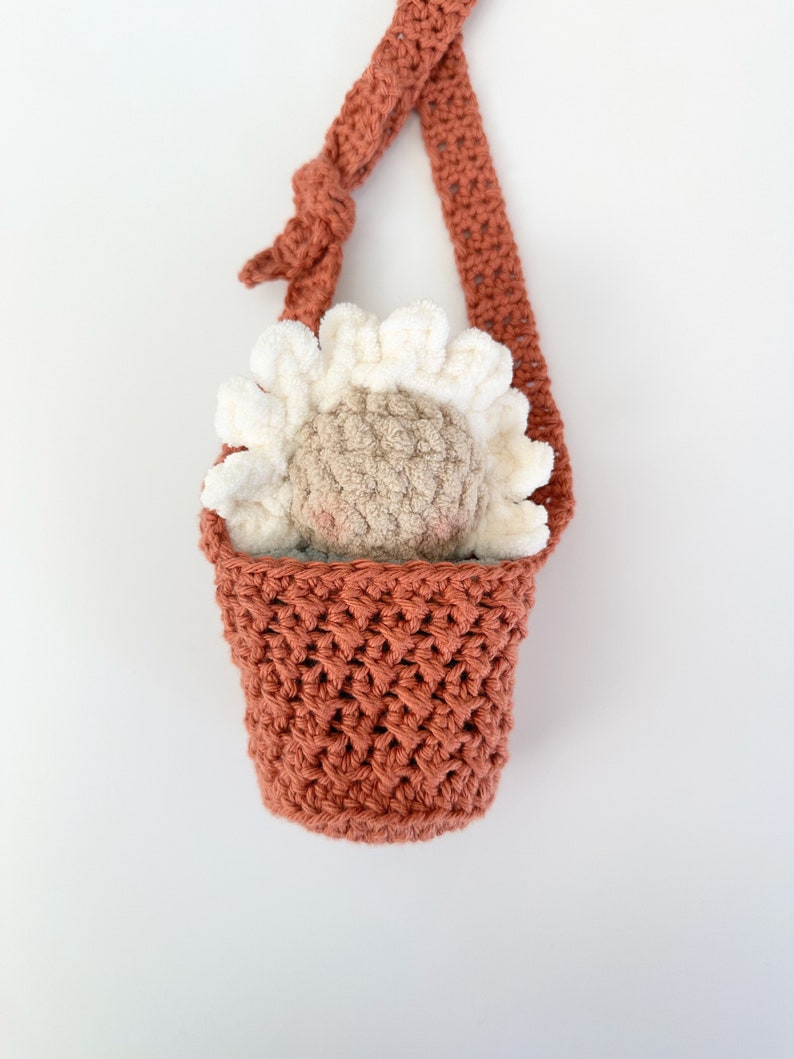 May include: A crocheted hanging planter with a terracotta colored pot and a white and beige crocheted flower inside.