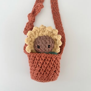 May include: A crocheted sunflower-shaped bag with a brown strap. The bag is filled with a brown and yellow crocheted sunflower toy.