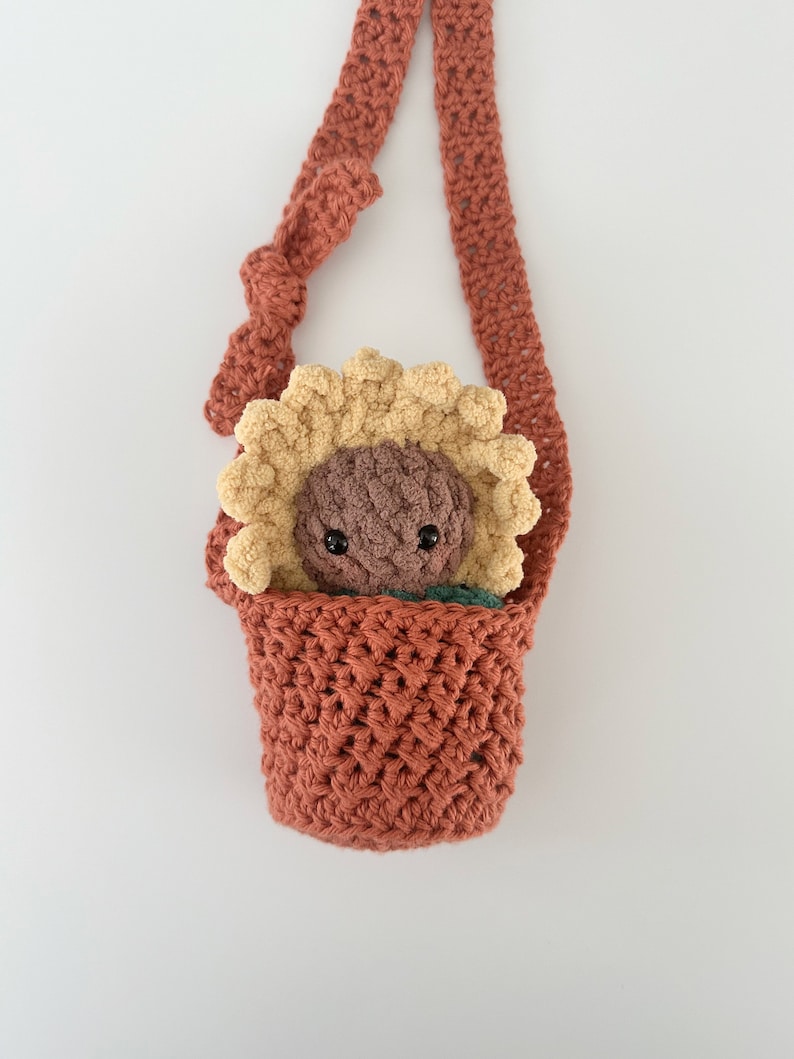 May include: A crocheted sunflower-shaped bag with a brown strap. The bag is filled with a brown and yellow crocheted sunflower toy.