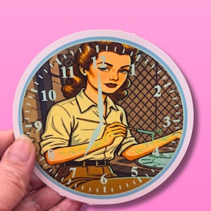 May include: A round clock sticker featuring a retro illustration of a woman painting. The clock face has a light blue border with black numbers and hands. The woman is wearing a yellow shirt and brown pants.
