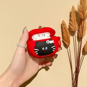 May include: A red apple-shaped AirPod case with a black cat face design. The case is open, revealing white AirPods. The cat has white whiskers and a white bow. The text "OHK" is on the side of the case.