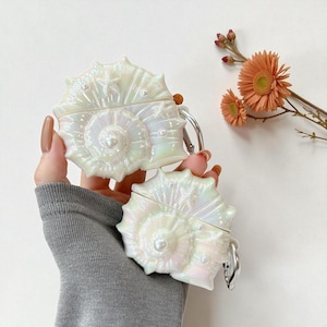 May include: Two iridescent white seashell-shaped cases with silver carabiner clips. The cases feature a spiral design with a central pearl. They are held in a hand. A sprig of orange flowers is in the background.