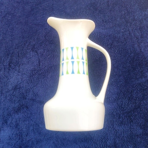 May include: A white ceramic pitcher featuring a flared spout and a curved handle. The body of the pitcher is decorated with a repeating pattern of vertical, hourglass-shaped motifs in alternating shades of light blue and lime green.