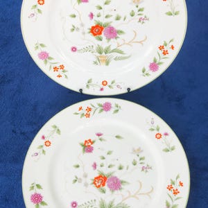 May include: Two round white ceramic dinner plates featuring a delicate floral pattern with pink, orange, and green botanical illustrations. The design includes clusters of small flowers and leaves arranged in a central bouquet and along the rim.