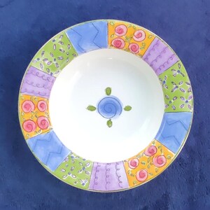 May include: A round ceramic bowl with a white center featuring a small blue spiral design surrounded by four green leaves. The wide rim is decorated with a colorful patchwork pattern of blue zigzags, orange circles, purple swirls, and green floral motifs.