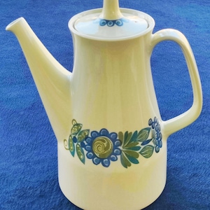 May include: A cream-colored ceramic coffee pot featuring a tall, tapered body, a long spout, and a handle. The pot is decorated with a floral pattern in shades of blue and green around the base and a matching circular design on the lid knob.