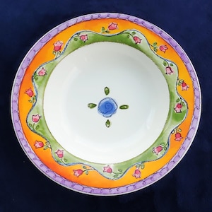 May include: A round ceramic bowl featuring a white center with a blue floral motif. The rim is decorated with a wavy green band adorned with small pink flowers, surrounded by a bright orange border and a purple scalloped edge pattern.