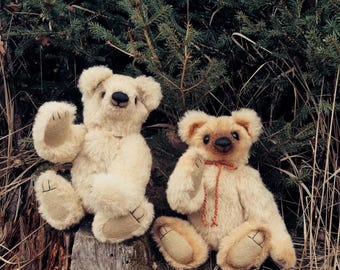 May include: Two plush teddy bears with cream-coloured fur, dark noses, and stitched paw pads. The bear on the right wears a thin, orange-brown cord tied in a bow around its neck. Both bears have rounded ears and are seated upright on a wooden surface.
