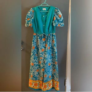 May include: A vintage teal dress with a floral pattern. The dress has a button-down front, short puff sleeves, and a drawstring waist. The floral pattern includes orange and white flowers on a teal background. The dress is hanging on a green hanger.
