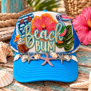 May include: A bright blue trucker hat featuring the embroidered text Beach Bum in gold glitter. The front panel displays a colorful hibiscus flower, a pelican, a flip-flop, and a coconut drink. A silver chain with starfish and seashells adorns the brim.