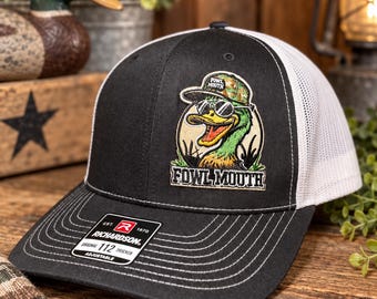 May include: A black and white trucker hat featuring a patch of a duck wearing sunglasses and a camouflage cap. The patch includes the text FOWL MOUTH. The hat has a black front panel, white mesh back, and a Richardson 112 sticker on the black brim.