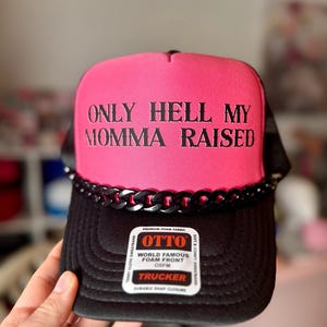 May include: A black and pink trucker hat with the text "ONLY HELL MY MOMMA RAISED" in black. The hat has a black chain detail and a white tag that reads "OTTO WORLD FAMOUS FOAM FRONT OSFM TRUCKER".