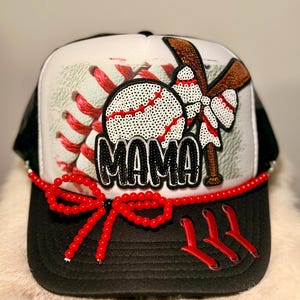 May include: A baseball-themed trucker hat with a black brim and a white and black mesh back. The front features a sequined baseball with red stitching, a brown bat, and the word "MAMA" in black sequins. A red beaded bow adorns the brim.
