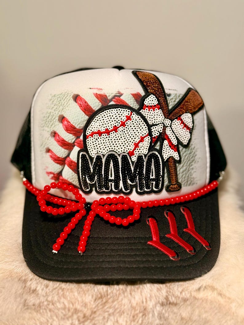 K&ouml;nnte beinhalten: Baseball-Trucker-Cap mit schwarzem Schirm und wei&szlig;-schwarzem Mesh-R&uuml;cken. Die Vorderseite zeigt einen paillettenbesetzten Baseball mit roten N&auml;hten, einen braunen Schl&auml;ger und das Wort "MAMA" in schwarzen Pailletten. Eine rote Perlen-Schleife verziert den Rand.