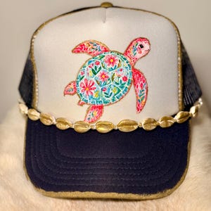 May include: A trucker hat with a navy blue brim and a white front panel. The front features a colorful embroidered sea turtle with floral details. The hat is trimmed with gold accents and adorned with gold-colored seashells.