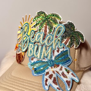 May include: Tan trucker hat with a sequined "Beach Bum" design. The design includes a sun, palm trees, and a bow with a leopard print pattern. The hat has a mesh back and a curved brim.