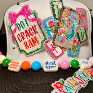 May include: A black and white trucker hat with colorful embroidered patches. The patches include the words "DOT CRACK BAM" and "MAHJO", along with images of flowers and a peacock. A beaded strand with colorful beads is attached to the hat.