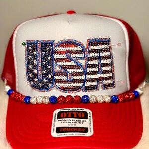 May include: A red and white trucker hat with the word "USA" in sequined letters. The hat features a red brim, a white front panel, and a red mesh back. A beaded embellishment in red, white, and blue decorates the brim.