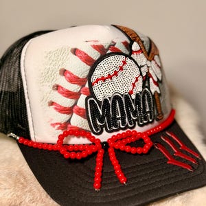 K&ouml;nnte beinhalten: Baseballkappe mit schwarzem Schirm und Mesh-R&uuml;ckseite. Die Vorderseite zeigt ein Baseball-Design mit dem Wort "MAMA" in schwarzen Pailletten. Eine rote Perlen-Schleife und Akzente erg&auml;nzen die Kappe.