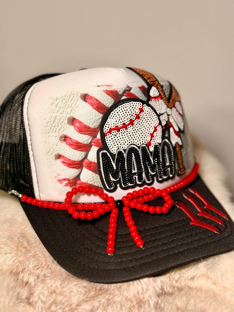 K&ouml;nnte beinhalten: Baseballkappe mit schwarzem Schirm und Mesh-R&uuml;ckseite. Die Vorderseite zeigt ein Baseball-Design mit dem Wort "MAMA" in schwarzen Pailletten. Eine rote Perlen-Schleife und Akzente erg&auml;nzen die Kappe.