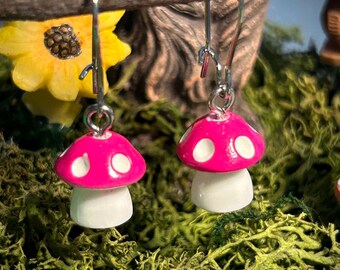 May include: A pair of whimsical mushroom earrings. Each earring features a bright pink cap with white spots atop a white stem. The earrings hang from silver-colored ear wires, set against a backdrop of green moss and a yellow sunflower.