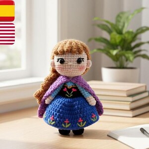 May include: A crocheted doll featuring a blue dress, purple cape, and brown hair. The doll has a detailed face and is positioned on a wooden surface. The doll is a character from a popular animated film.