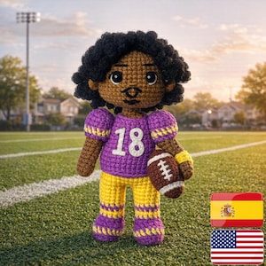 Könnte beinhalten: Gehäkelte Puppe eines Footballspielers, trägt ein lila Trikot mit der Nummer 18 und gelbe Hosen. Die Puppe hat schwarze Haare und hält einen braunen Football. Die Puppe steht auf einem grünen Feld.