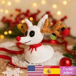May include: A white crocheted reindeer with brown antlers, a red nose, and a red scarf. The reindeer has black eyes and is decorated with small, colorful ornaments. The image includes the text "Crochet pattern".