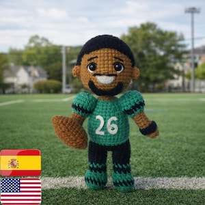 Könnte beinhalten: Handgefertigte Häkelpuppe eines Footballspielers. Sie trägt ein grünes Trikot mit der Nummer 26, eine schwarze Hose und hält einen braunen Football. Die Puppe hat ein lächelndes Gesicht und steht auf einem grünen Feld.
