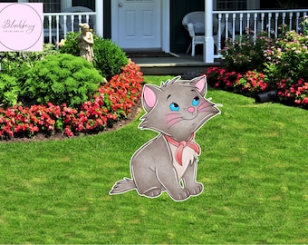 Op de afbeelding: Een cartoon kat tuinornament. De kat is grijs met roze oren en een roze strik. Het heeft blauwe ogen en roze wangen. De kat zit op groen gras voor een huis met een witte hek en rode bloemen.