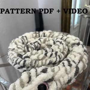 May include: Crocheted snake toy with a white and gray striped pattern. The snake has a black eye with a pink glitter accent. The image includes the text "PATTERN PDF + VIDEO" and "ERENA'S CROCHET STUDIO".