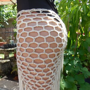 May include: A beige crochet skirt with a geometric net pattern and fringe detail. The skirt is made of a light-colored yarn and is worn over a black swimsuit. The skirt is designed to be worn at the waist.