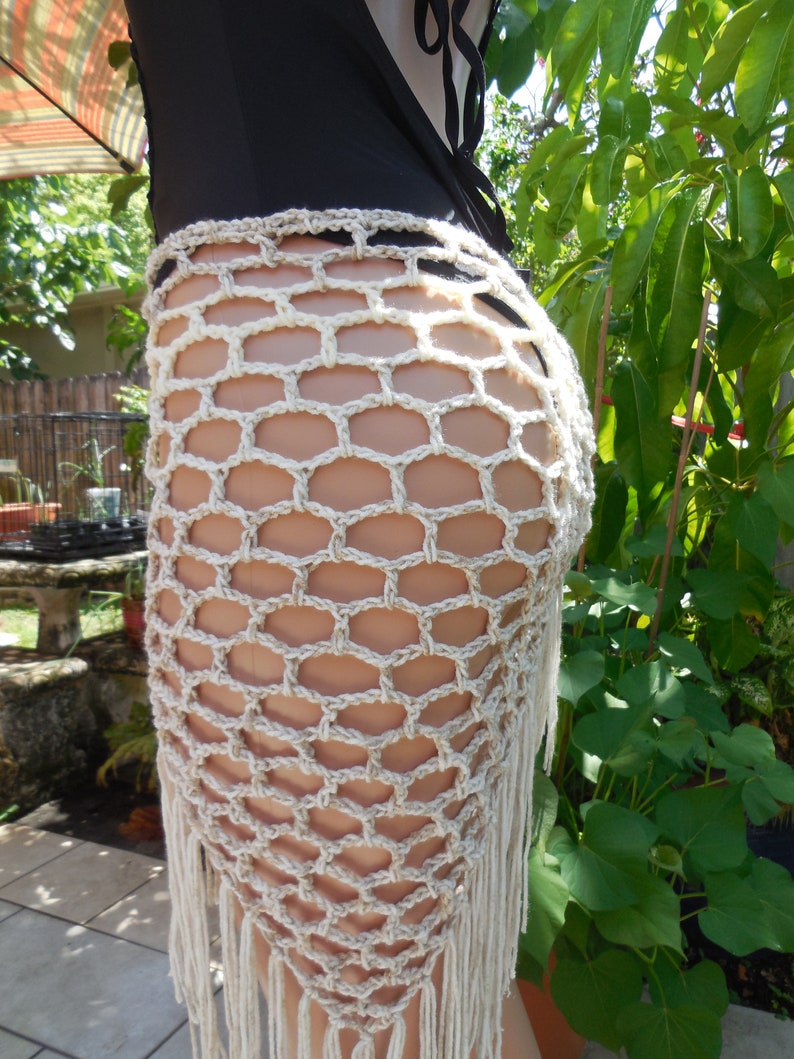 May include: A beige crochet skirt with a geometric net pattern and fringe detail. The skirt is made of a light-colored yarn and is worn over a black swimsuit. The skirt is designed to be worn at the waist.