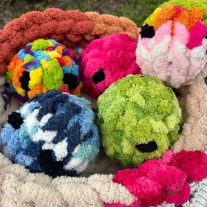 May include: A collection of colorful, handmade crochet balls in a white basket. The balls are various colors, including pink, green, blue, and rainbow. Each ball has a black accent. The basket is surrounded by a thick, braided yarn in shades of beige, pink, and orange.