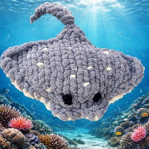 Pode incluir: Um brinquedo de pelúcia de arraia cinzenta, feito à mão, com manchas brancas e olhos pretos. A pelúcia tem uma cauda curva e uma parte inferior branca. O fundo é uma cena subaquática com corais e peixes.