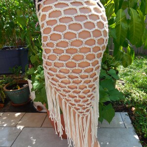 May include: A beige crochet skirt with a honeycomb pattern and fringe detail. The skirt is handmade and has a unique, open design. The fringe hangs down from the bottom edge of the skirt.