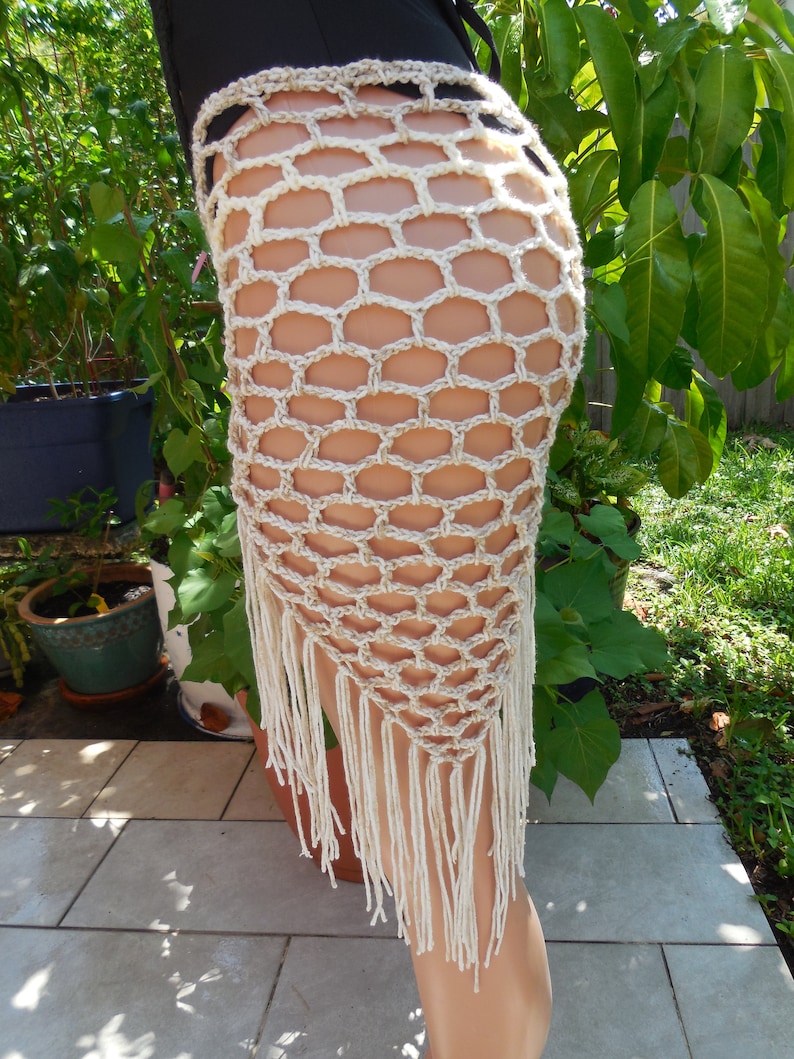 May include: A beige crochet skirt with a honeycomb pattern and fringe detail. The skirt is handmade and has a unique, open design. The fringe hangs down from the bottom edge of the skirt.