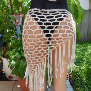 May include: A beige crochet skirt with a fringe hem. The skirt is made of a net-like pattern and is worn over a black swimsuit. The skirt is a unique beach cover-up.