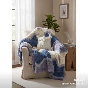 May include: A cozy armchair draped with a chunky knit blanket in shades of blue, lavender, and cream. A matching decorative pillow with a textured design rests on the chair. The room has a neutral color palette with a plant and artwork.