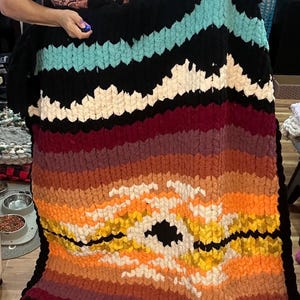 May include: A large, handmade blanket with a vibrant, sunset-inspired design. The blanket features a black border, turquoise, white, burgundy, orange, and yellow stripes. The blanket is made of a thick, textured yarn.